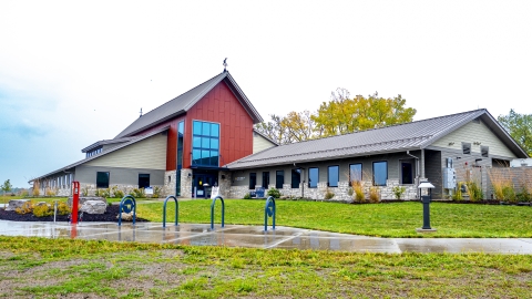 Photo is taken from the ground looking up on a new facility with stone and red siding under a blue and white sky with green grass in foreground