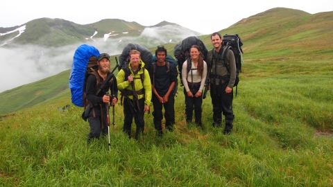 Five young people with big backpacks stand in a meadow with snow-covered mountains behind them.