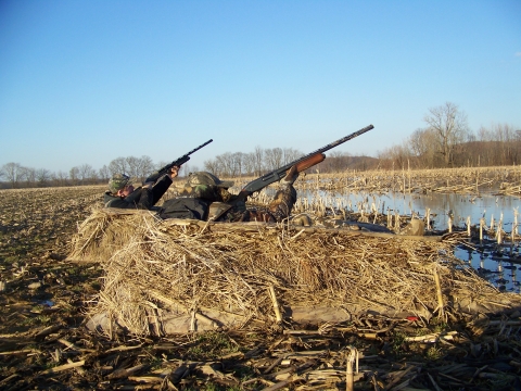 Two hunters in dirt raised camouflaged dirt bunks with shotgun raised. 