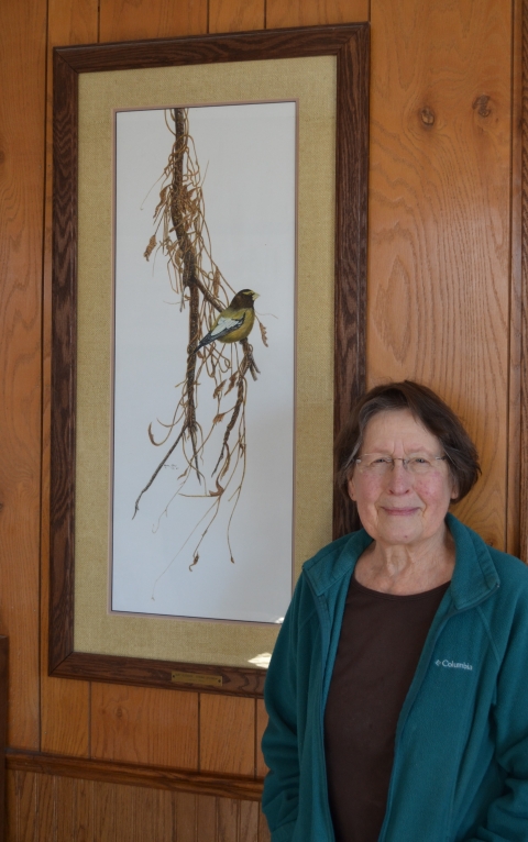 Artist Marion Otnes stands next to her framed and matted watercolor of an evening grosbeak