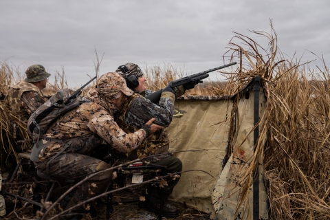 Person is camouflage hunting gear crouches behind a second person in a wheelchair in a duck hunting blind