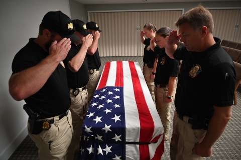 The honor guard stands over a coffin draped in an American flag. Each of them is saluting and looking down at the coffin/flag.