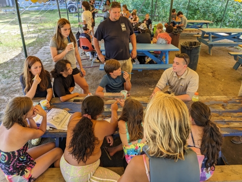 a group of kids and a Service employee sit and talk at a picnic bench