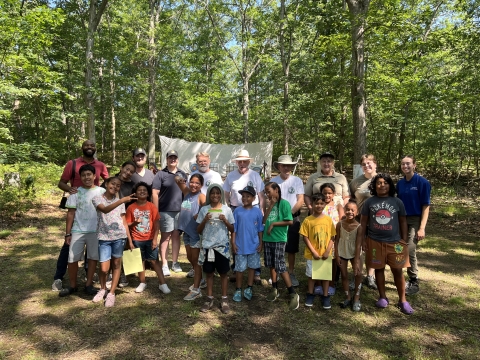 a large group of kids and adults smile in front of an archery range in the forest