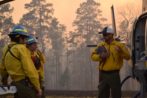 Four firefighters stand next to a helicopter talking. The background is smoky with large trees.