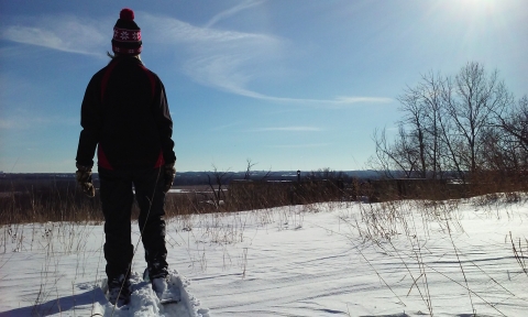 A person wearing snowshoes and a winter hat with a red pompom faces away from the camera as they look out across the Minnesota River Valley from the field next to the overlook behind the Bloomington Education and Visitor Center.