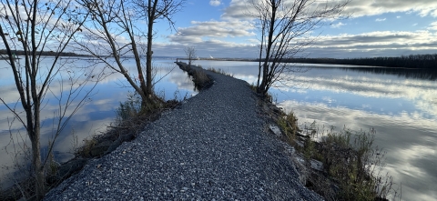 a stone jetty with a few sparse trees cuts through a still lake reflecting blue skies and clouds
