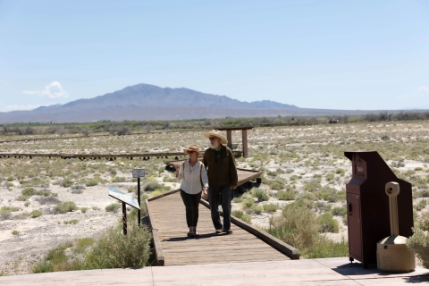 Two people walk down a raised boardwalk in a desert environment.