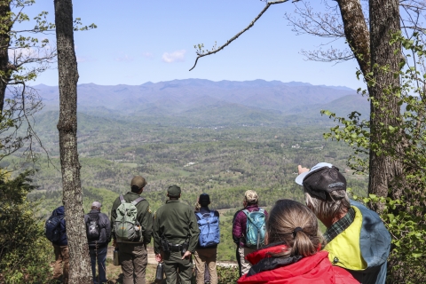 Several people standing on the peak of a low mountain, looking out across a valley to a distant mountain range