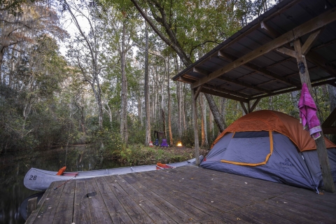 photo of wooden shelter with a portion under a roof with a gray and orange tent and canoe in foreground. In background is a campfire with trees all around.