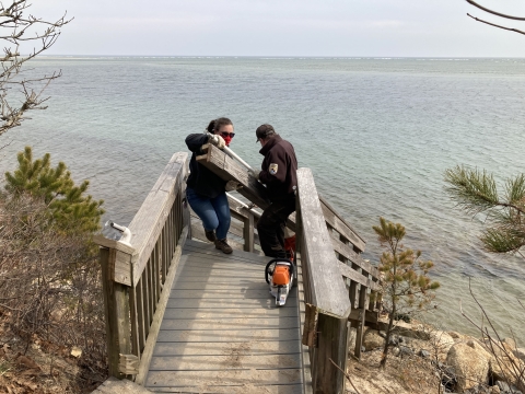 two people hold large planks of wood together while walking up a set of stairs leading to a beach below.