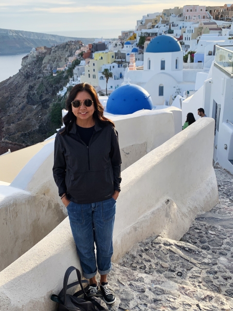 Susan Machida stands along cobblestone steps. The buildings of Santorini, Greece, adorn the background. Their ivory white colors contrast against the brown cliffsides.