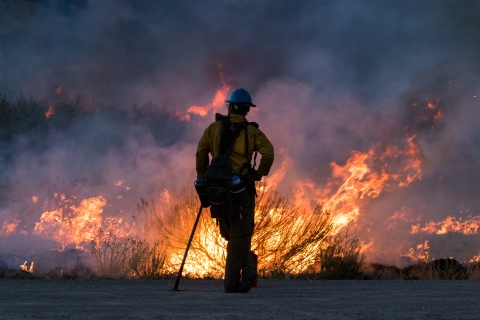 A firefighter leans against a fire tool looking out at a fire at night.