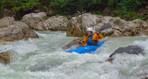A woman in a blue kayak paddles down white water. She wears a white helmet, orange jacket, and blue bib.