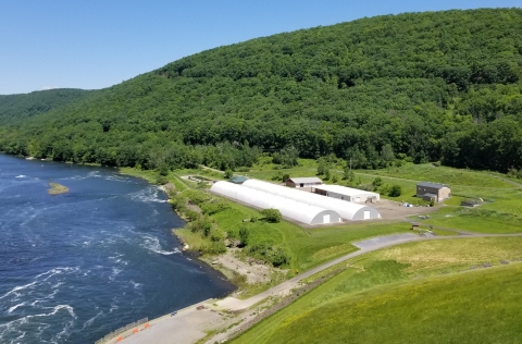 A drone view of two quonset building that are very long and bright white against a very green landscape. One one side is a tree covered hill and on the other is a large body of water.