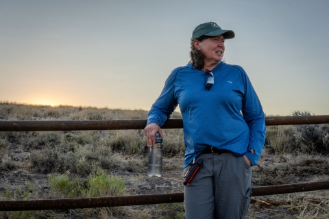 Fish and Wildlife Service employee stands in front of a fence line with sagebrush in the background.