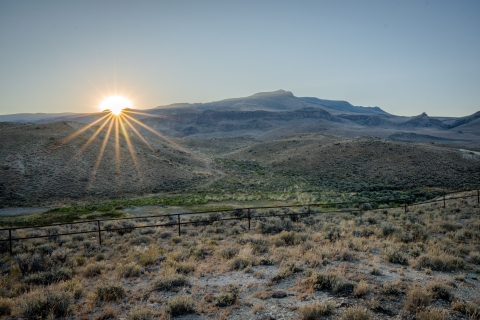A sagebrush landscape at sunrise. A fence runs through the foreground and there are mountains stretching into the distance.