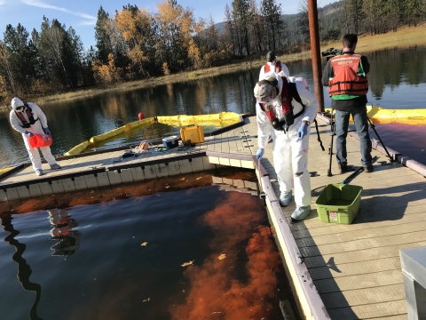 Three individuals in full protective gear spray an orange chemical into a lake while another individual sets up a camera
