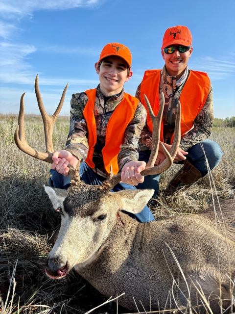 Two young brothers wearing orange and camo with the younger one holding a harvest mule deer by the antlers