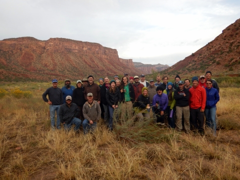 A group of individuals pose for a group photo in a grassland with a backdrop of canyons