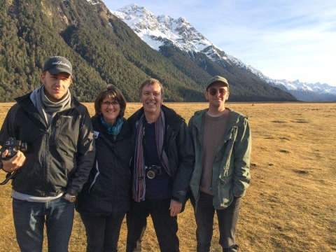 A group of four people stand in an outdoor expanse of brown grass and large mountains covered with trees and snow