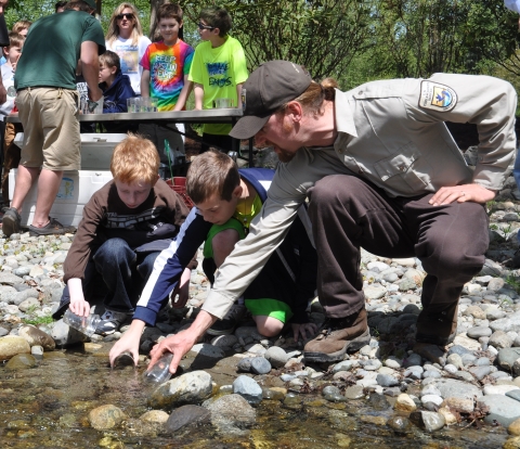 Man in Service uniform next to two boys on creek bank releasing juvenile salmon from cups. Youth and adults in the background.