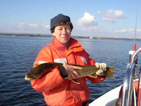 a woman in a boat wearing an orange coat and navy blue hat holds a large fish across her body, with water behind