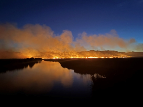 Flames glow in front of dark mountains and river. 
