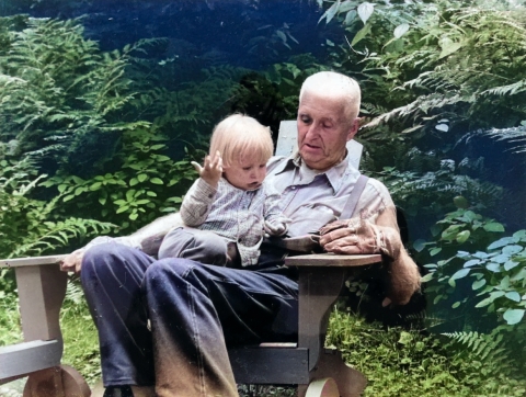 Colorized photo of an elderly man in jeans, suspenders, and leather work gloves sitting on an Adirondack-style outdoor chair with a small boy on his lap, with a background of ferns and shrubs.