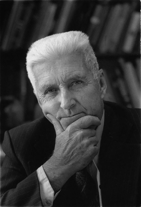 Black and white portrait of an older man with crew-cut white hair and a dark suit, leaning his chin on his hand, looking straight into the camera with a slight smile, bookshelves in the background.