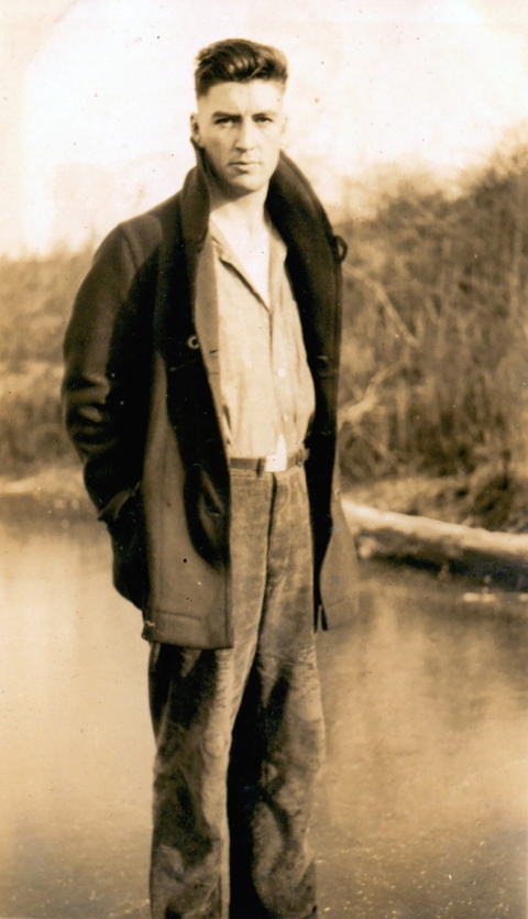 Sepia photo of a young man in an open coat, standing by water with a high bank on the opposite side.