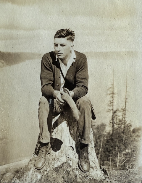 Sepia photo of a young man in sweater, collared shirt, trousers, and leather shoes sitting on a stump on a high place with a background overlooking trees below, water, and distant hills.
