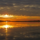 A gold and orange sunset reflects on the still waters of Back Bay. A line of trees denotes the horizon.