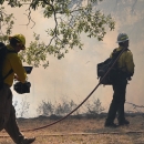 A cameraman follows a firefighter who is walking toward small flames and a smoky forested landscape. The firefighter has a hose slung over his shoulder and is dragging the rest on the ground.