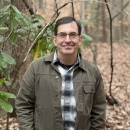Robert Tawes is standing and wearing a green jacket , jeans, and glasses with trees and brown leaves in the background