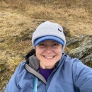 A person wearing a visored hat under a knit cap and a rain jacket takes a selfie with tundra and rocks behind them.