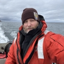 A USFWS staff member in a high visibility float coat personal floatation device and a USFWS Refuge uniform beanie takes a selfie near the stern of a boat. Open water in the background. Some land on the horizon can be seen. 