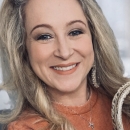 Karman has blond hair is wearing an orange top. She has a beautiful smile.