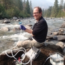 A smiling man in glasses holds scientific equipment while sitting on rocks by a stream.