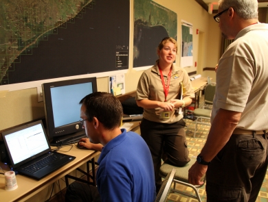 Three people are busy at work in the Operations Center during the aftermath of the Deepwater Horizon oil spill. One person is focused on a computer screen, with a map of the impacted area over their head. The two other people are talking to each other.