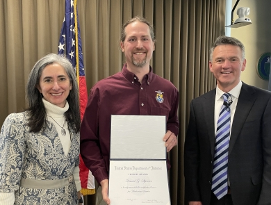 Three people smile standing in front of an American flag, and the man in the middle holds an award