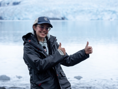 A young woman in a dark rain jacket smiles and holds up two thumbs up. She stands in front of icy water, with a large glacier visible in the background.