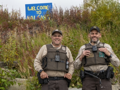 Two uniformed Federal Wildlife Officers pose for a picture with a painted sign in the background on a hill reading "welcome to Kalskag" 