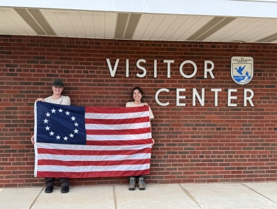 2 people hold up Betsy Ross flag in front of brick wall saying Visitor Center