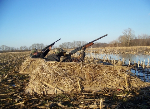 Two hunters in dirt camouflage bunker with shot guns raised to the sky.
