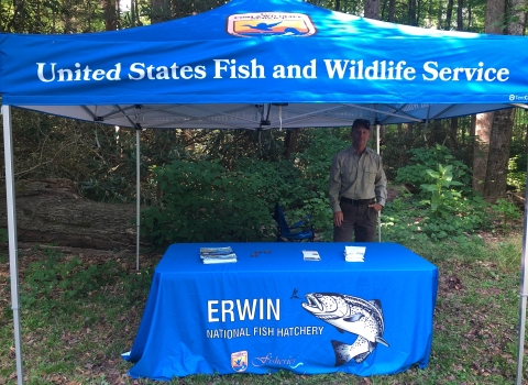 USFWS employee pictured standing behind a table with a blue tablecloth with the Erwin NFH logo and under a blue pop up tent with USFWS logo.