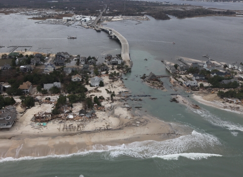 image shows an island neighborhood and houses connected to mainland by a bridge, which has been washed out by water. Debris litters the shoreline