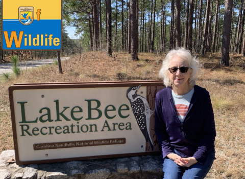 Deb Adams sits on the welcome sign at Lake Bee Recreation area at Carolina Sandhills National Wildlife Refuge. The sign has a woodpecker carved into it. Deb is wearing sunglasses and smiling. The WoW: Woman of Wildlife logo is in the upper left corner.