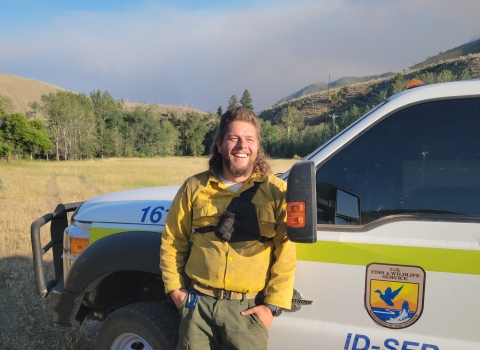 A man in fire clothes- a yellow shirt and green pants- is leaning against a truck with a FWS logo on the door. The man is smiling.