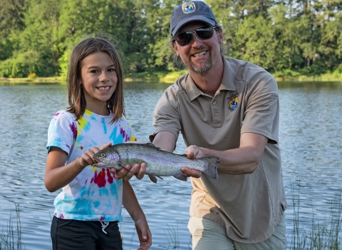 Male Service employee stands beside Big Brothers Big Sisters event participant as he holds a rainbow trout she caught. Lake in background.